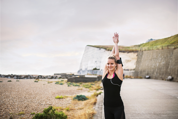 Young sporty woman runner with earphones and smartphone in an armband outside by the beach, stretching.