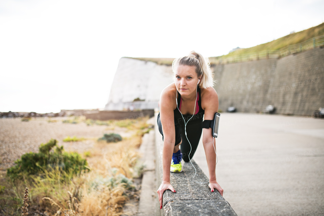 Young sporty woman runner with earphones and smartphone in an armband outside in nature, stretching.