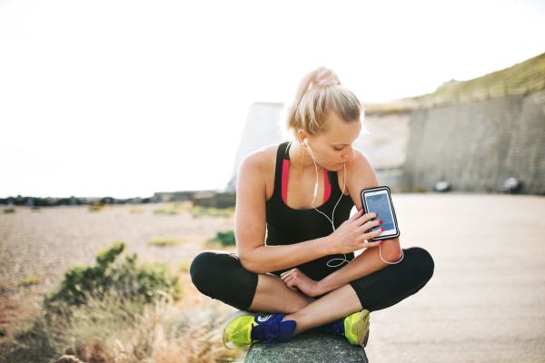 Young sporty woman runner with earphones sitting outside on the beach in nature, using smartphone in armband.