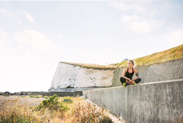 Young sporty woman runner with earphones and smartphone in armband sitting outside on the beach in nature, listening to music and resting.