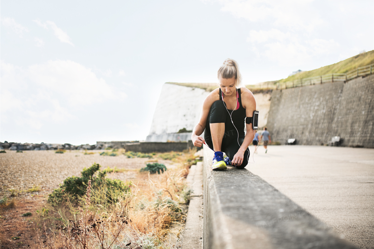 Young sporty woman runner with earphones and smartphone in an armband outside by the beach, tying shoelaces.