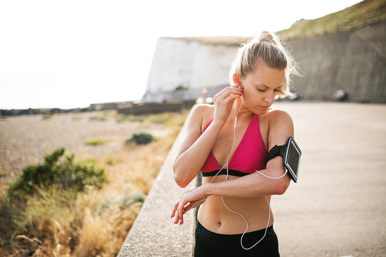 Young sporty woman runner with earphones and smartphone in armband standing outside on the beach in nature, listening to music and resting.