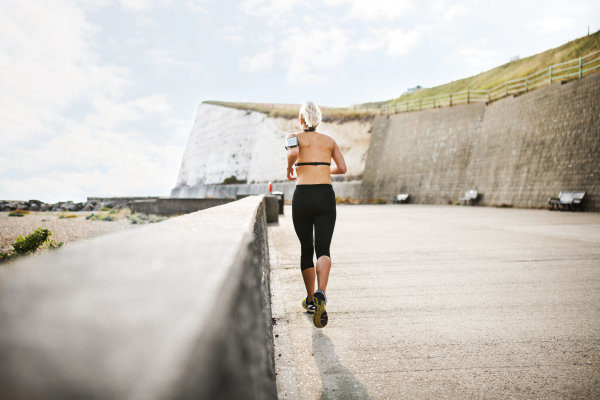 A rear view of young sporty woman runner with smartphone in an armband running outside on a beach in nature.