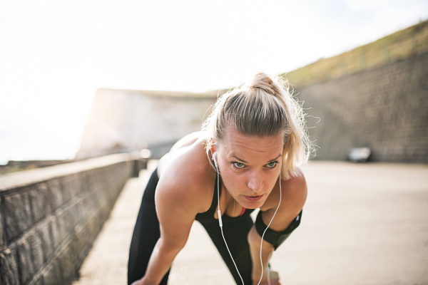 Young sporty woman runner with earphones and smartphone in an armband outside by the sea, resting.