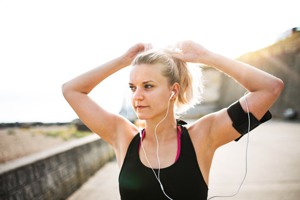 Young sporty woman runner with earphones and smartphone in armband standing outside on the beach in nature, listening to music and resting.