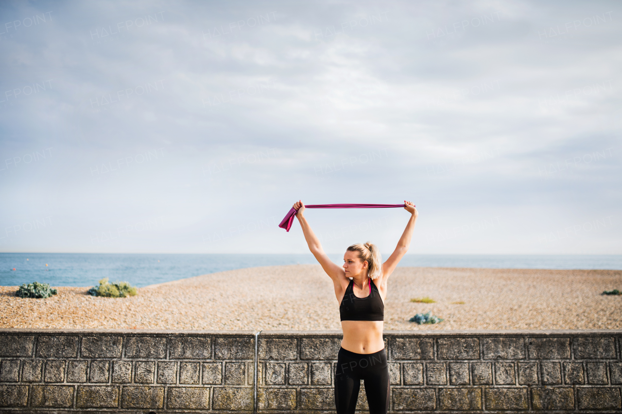 Young sporty woman runner doing exercise with elastic rubber bands outside on a beach in nature.