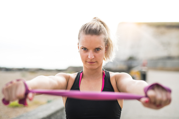 Young sporty woman runner doing exercise with elastic rubber bands outside on a beach in nature.