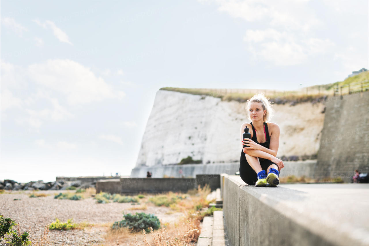 Young sporty woman runner with water bottle sitting outside on the beach in nature, resting. Copy space.