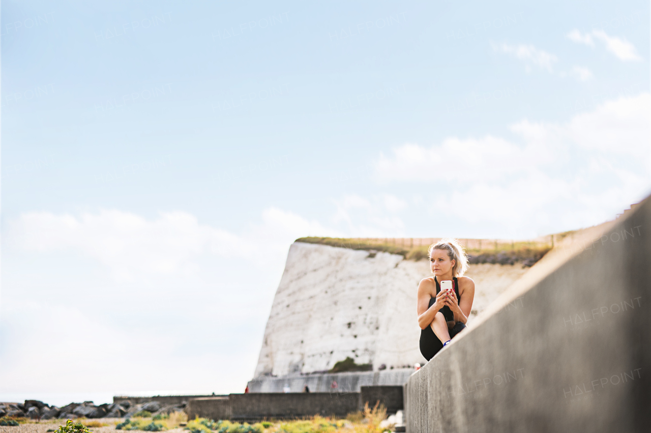 Young sporty woman runner with earphones and smartphone sitting outside on the beach in nature, listening to music and resting. Copy space.