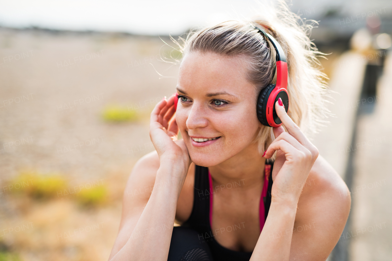 A close-up of young woman runner with black and red headphones resting outside by the sea, listening to music.