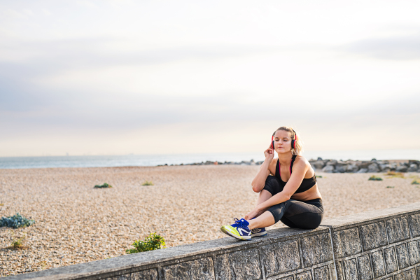 A young woman runner with black and red headphones resting outside by the sea, listening to music.