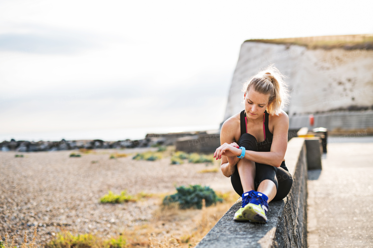 Young sporty woman runner resting outside in nature, sitting on a wall and setting smartwatch.