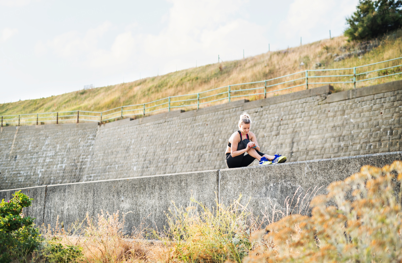 Young sporty woman runner sitting on a concrete wall outside, using smartwatch and resting.