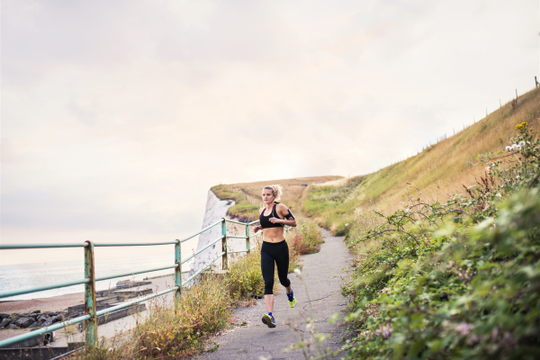 Young sporty woman runner with earphones running by the sea outside in nature, listening to music.