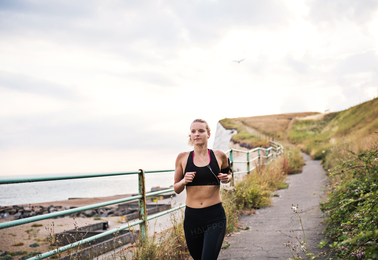 Young sporty woman runner with earphones running by the sea outside in nature, listening to music.