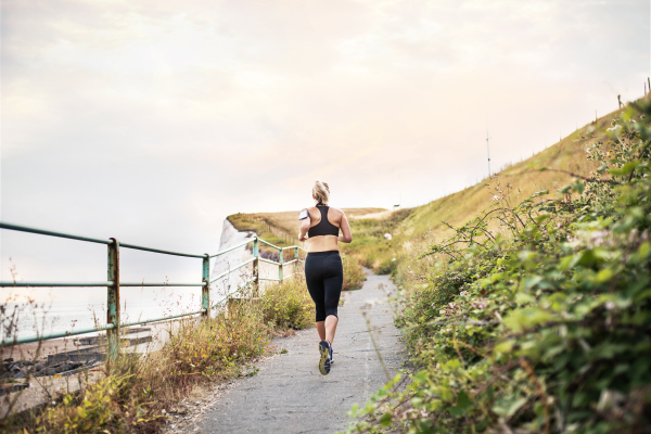 A rear view of young sporty woman runner with smartphone in an armband running by sea in nature.