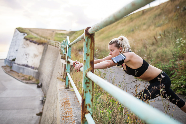 Young sporty woman runner with earphones and smartphone in armband stretching outside on the beach in nature, listening to music.