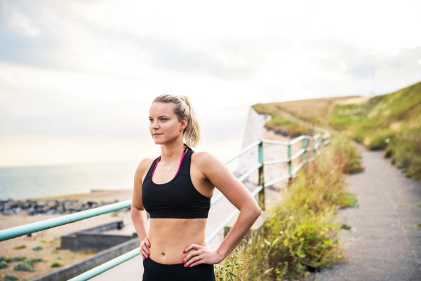 Young sporty woman runner in black activewear standing outside by the seaside, resting. Copy space.