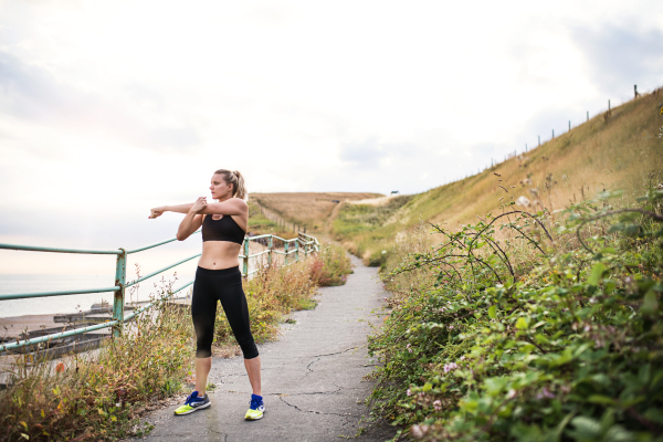 Young sporty woman runner in black activewear standing outside in nature, stretching. Copy space.