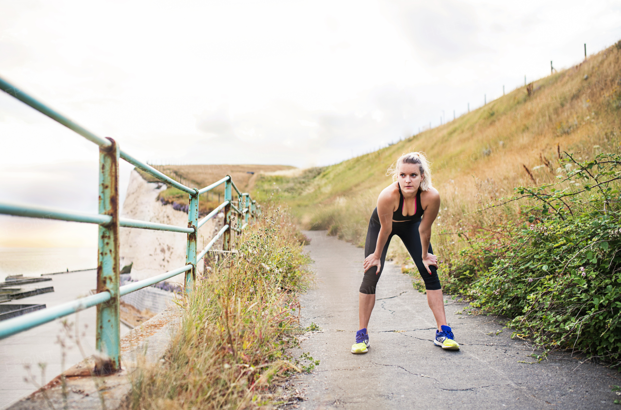 Young sporty woman runner in black activewear standing outside by the seaside, resting. Copy space.