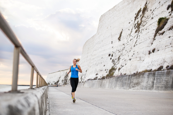 Young sporty woman runner with earphones running outside on the beach in nature, listening to music. Copy space.