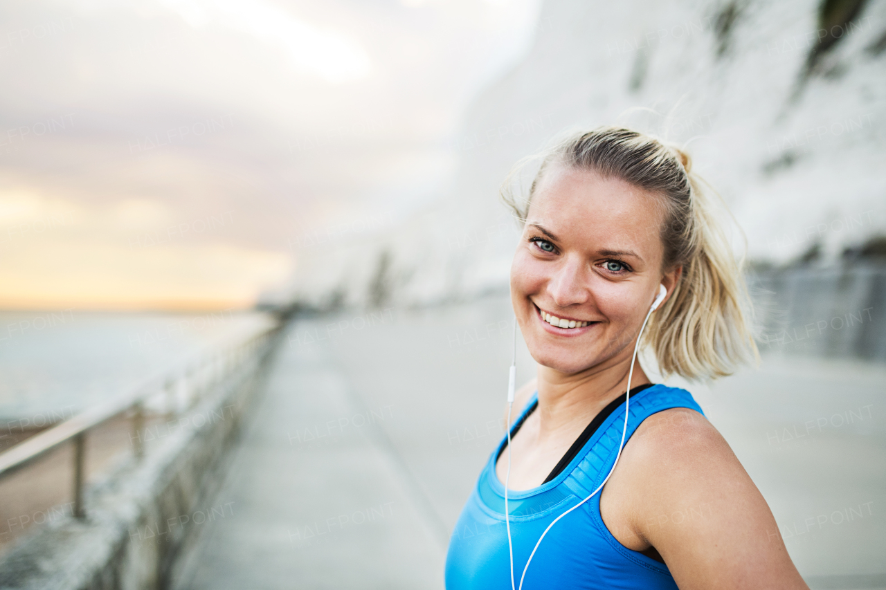 Young sporty woman runner with earphones standing outside on the beach in nature, listening to music and resting. Copy space.