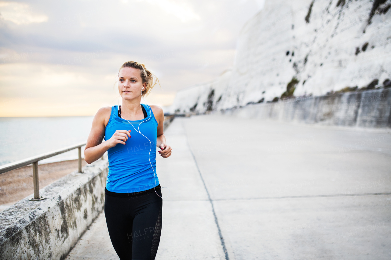 Young sporty woman runner with earphones running outside on the beach in nature, listening to music. Copy space.