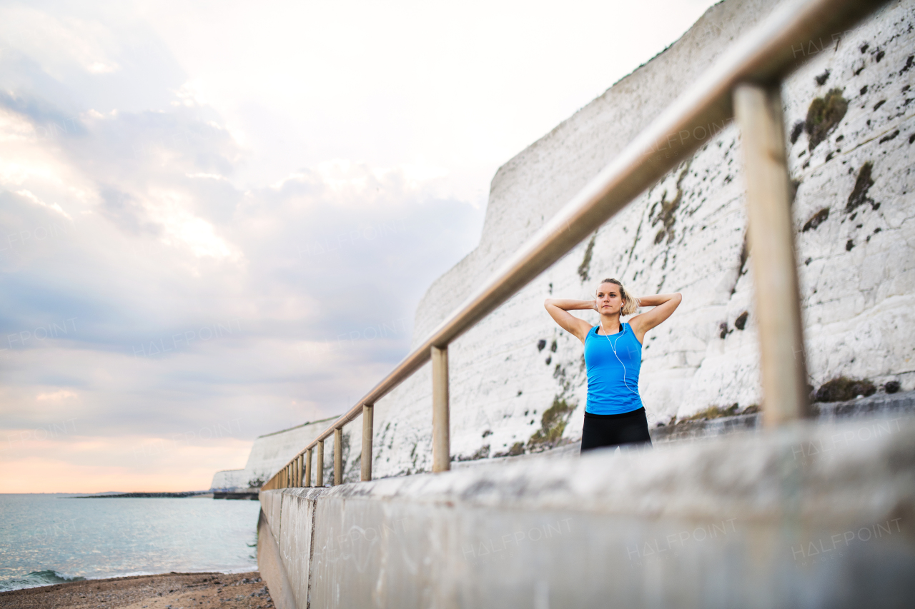 Young sporty woman runner with earphones stretching outside on the beach in nature. Copy space.