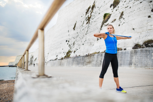 Young sporty woman runner doing exercise with elastic rubber bands outside on a beach in nature. Copy space.