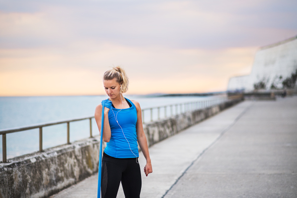 Young sporty woman runner doing exercise with elastic rubber bands outside on a beach in nature. Copy space.