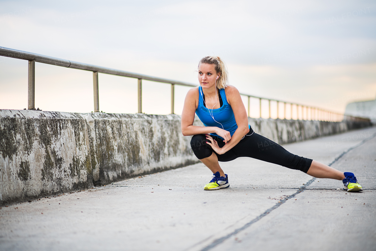 Young sporty woman runner with earphones stretching outside by the sea in nature. Copy space.