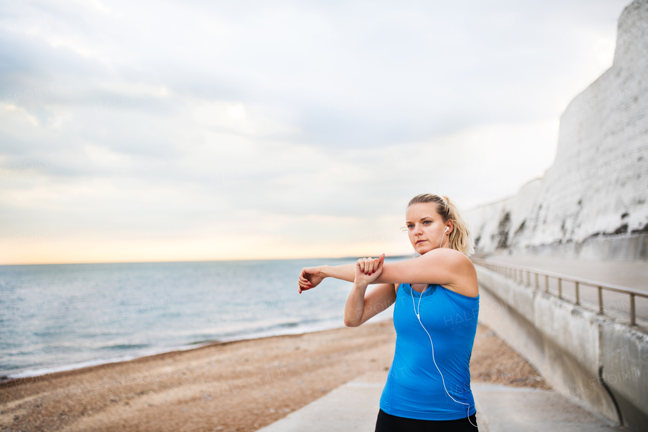 Young sporty woman runner with earphones standing outside on the beach in nature, listening to music and stretching. Copy space.