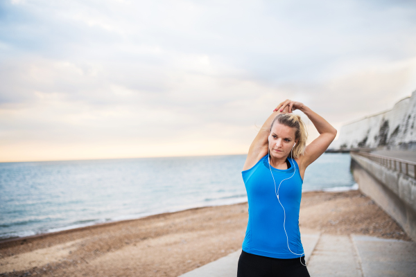 Young sporty woman runner with earphones standing outside on the beach in nature, listening to music and stretching. Copy space.