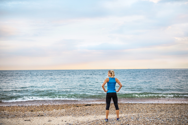 Young sporty woman runner in blue sportswear standing outside on the beach in nature, resting. Rear view.