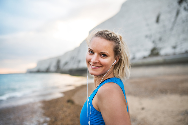 Young sporty woman runner with earphones standing outside on the beach in nature, listening to music and resting. Copy space.