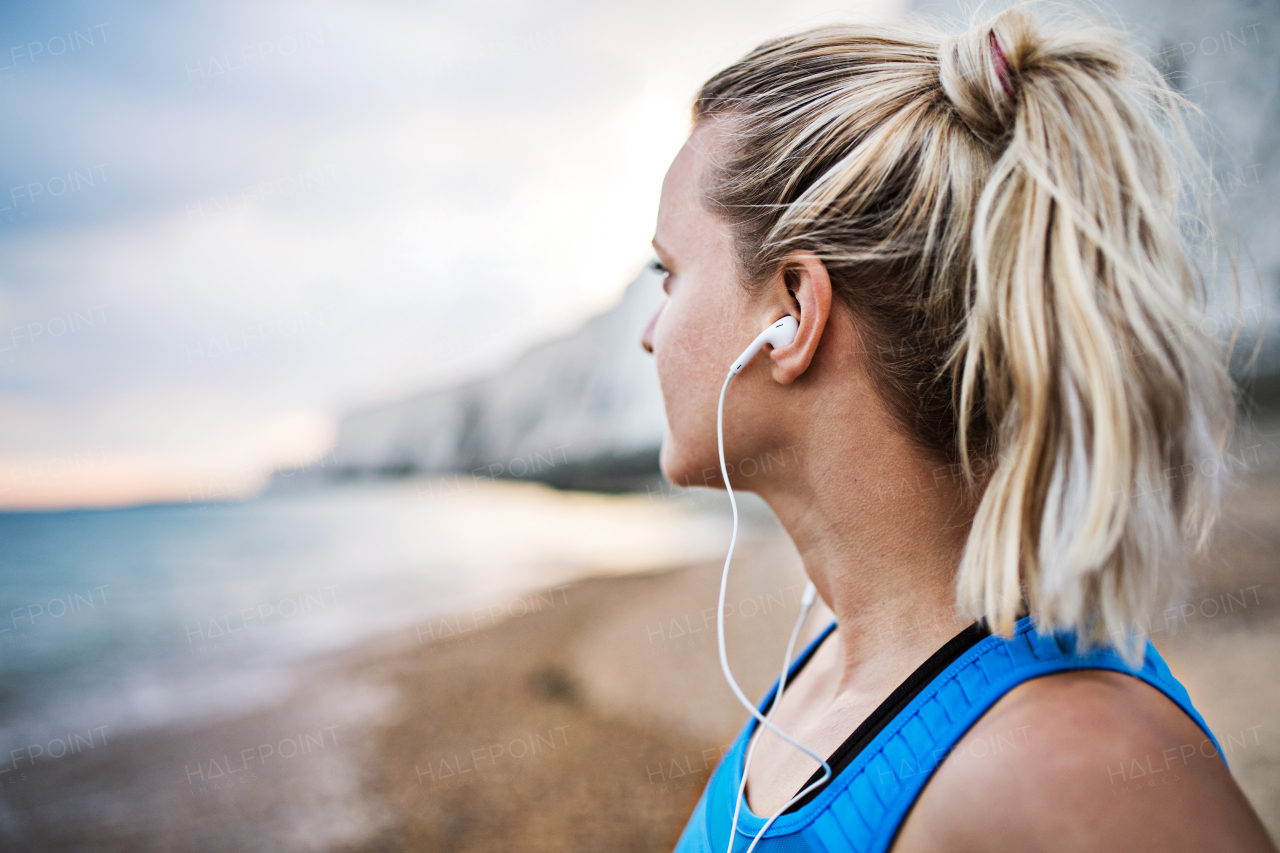 Young sporty woman runner with earphones standing outside on the beach in nature, listening to music and resting. Copy space.