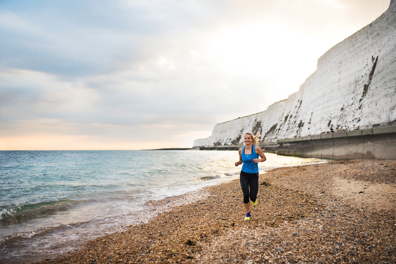 Young sporty woman runner with earphones running outside on the beach in nature, listening to music. Copy space.