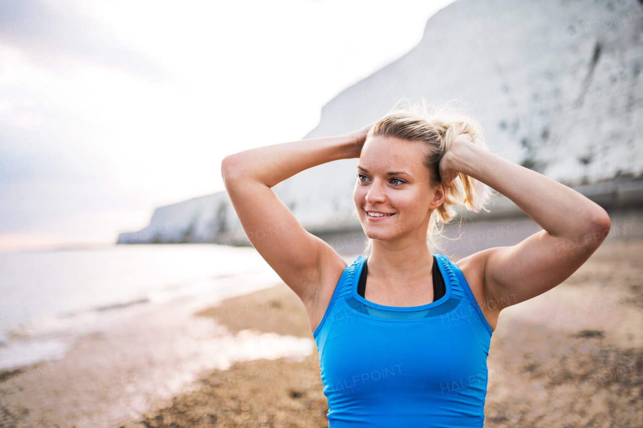 Young sporty woman runner in blue sportswear standing outside on the beach in nature, resting. Copy space.