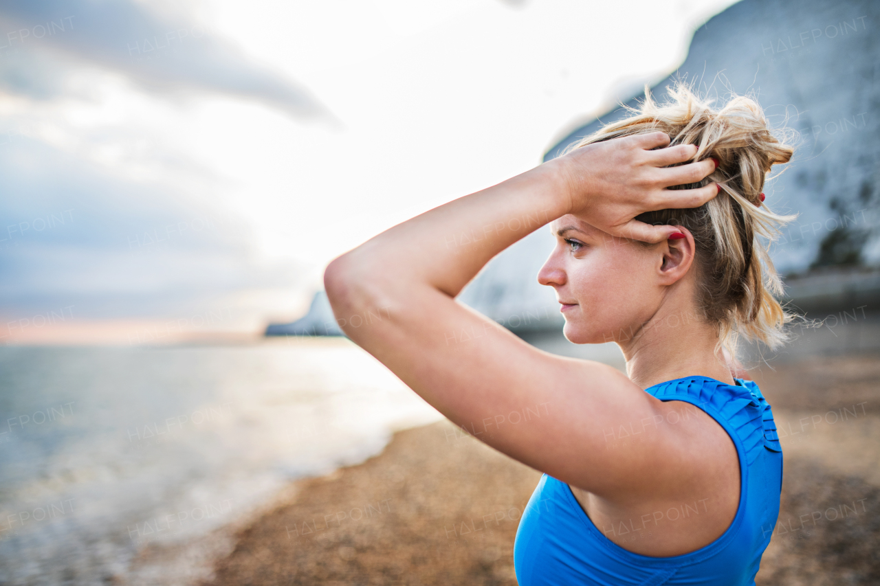 Young sporty woman runner in blue sportswear standing outside on the beach in nature, resting. Copy space.