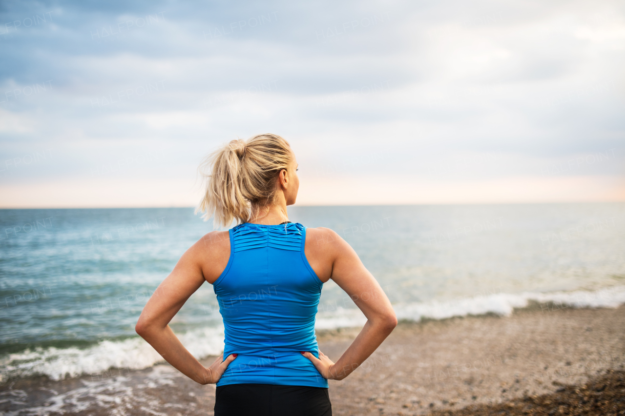 Young sporty woman runner in blue sportswear standing outside on the beach in nature, resting. Rear view.