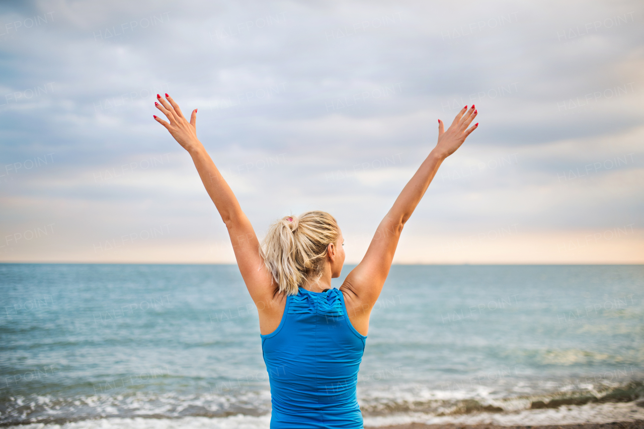 Young sporty woman runner in blue sportswear standing outside on the beach in nature, resting. Rear view.