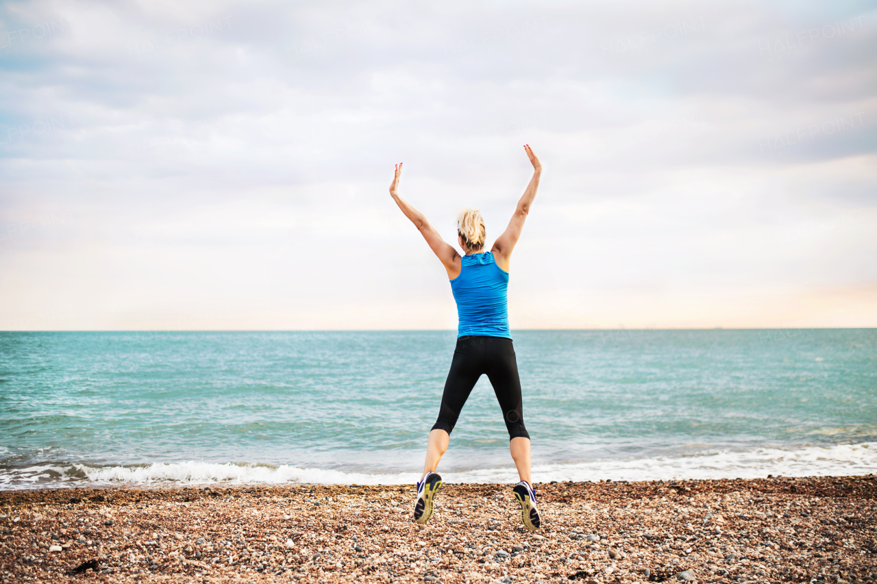 Young sporty woman runner in blue sportswear jumping outside on the beach in nature. Rear view.