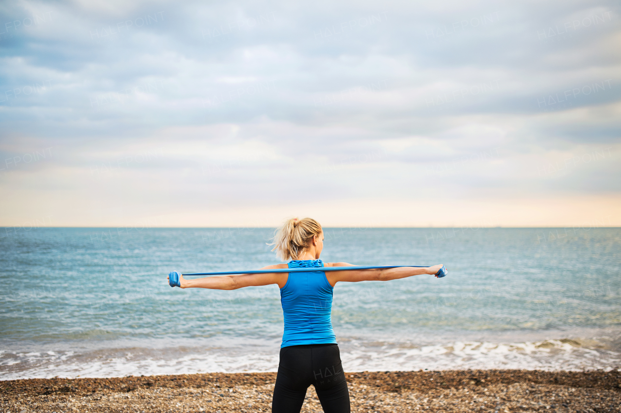 Young sporty woman runner doing exercise with elastic rubber bands outside on a beach in nature. Rear view.