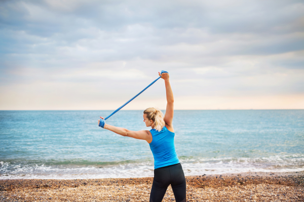 A rear view of young sporty woman runner doing exercise with elastic rubber bands outside on a beach in nature.