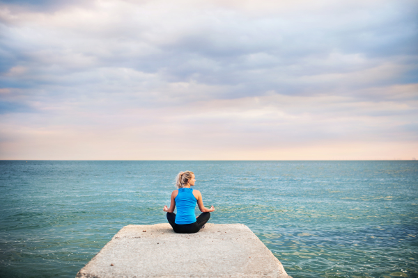 Rear view of a young sporty woman sitting on a pier, doing yoga exercise by the ocean outside. Copy space.