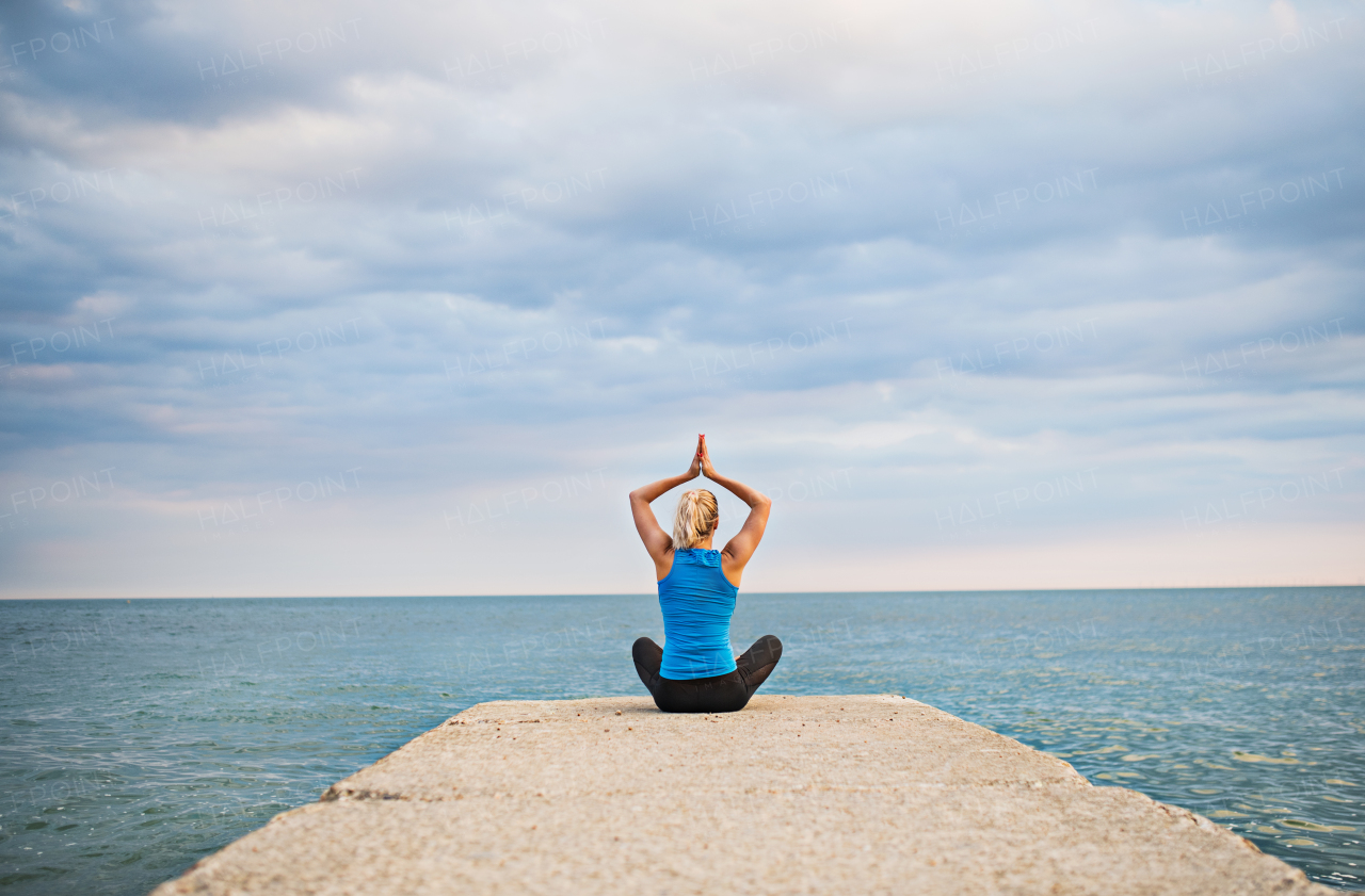 Rear view of a young sporty woman sitting on a pier, doing yoga exercise by the ocean outside. Copy space.