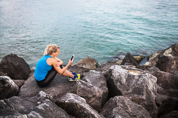 Young sporty woman runner with earphones and smartphone sitting on rocks by sea, listening to music.