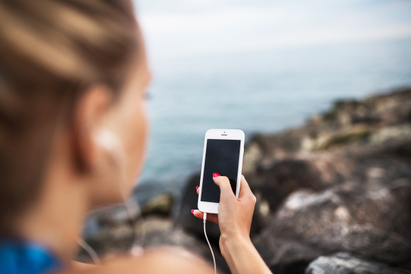 Unrecognizable young sporty woman runner with earphones and smartphone sitting on rocks by sea, listening to music. Close-up.