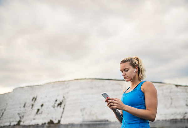 Young sporty woman runner with earphones and smartphone standing outside on the beach in nature, listening to music and resting. Copy space.