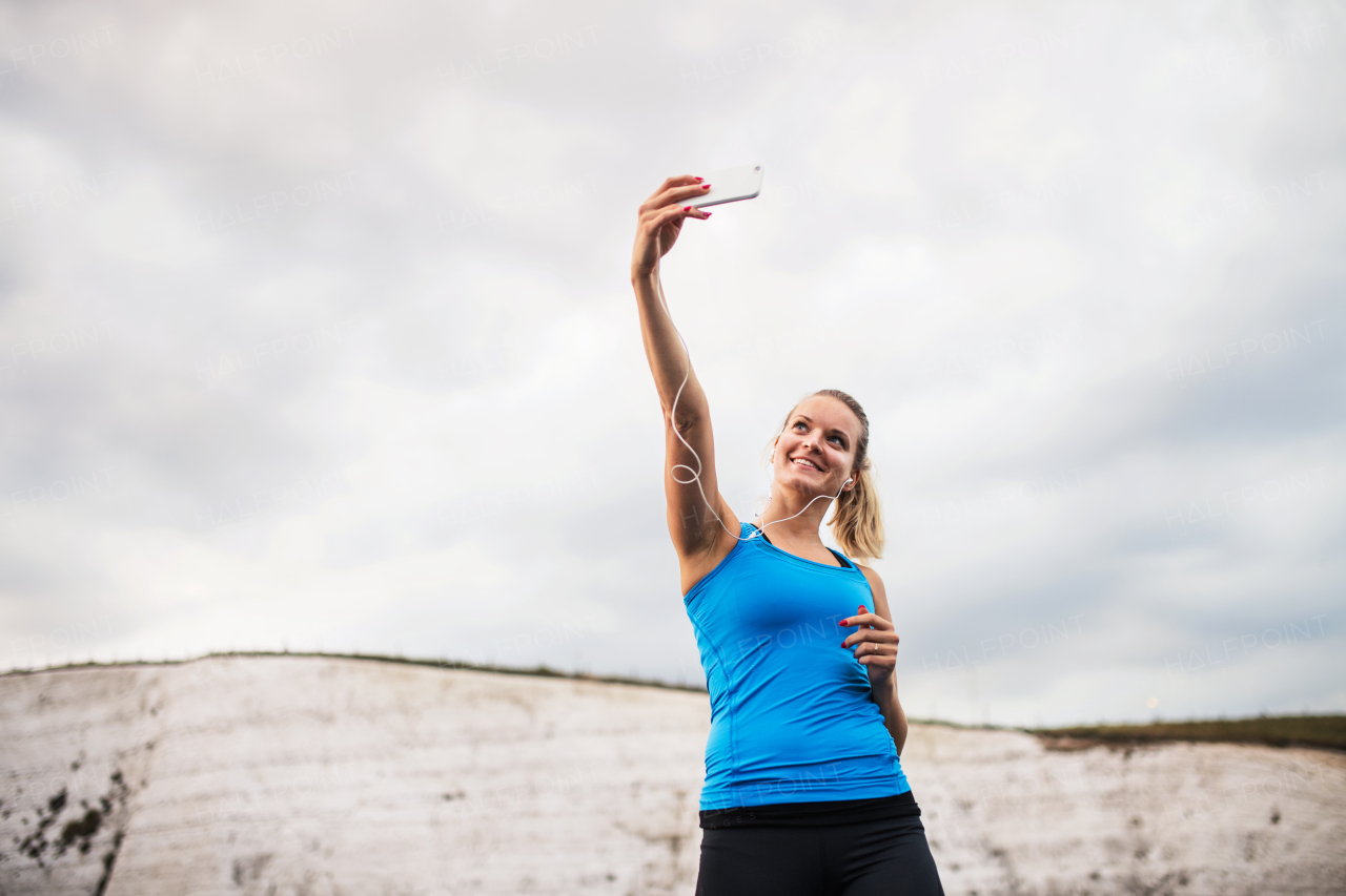 Young sporty woman runner with earphones and smartphone on the beach, taking selfie. Copy space.